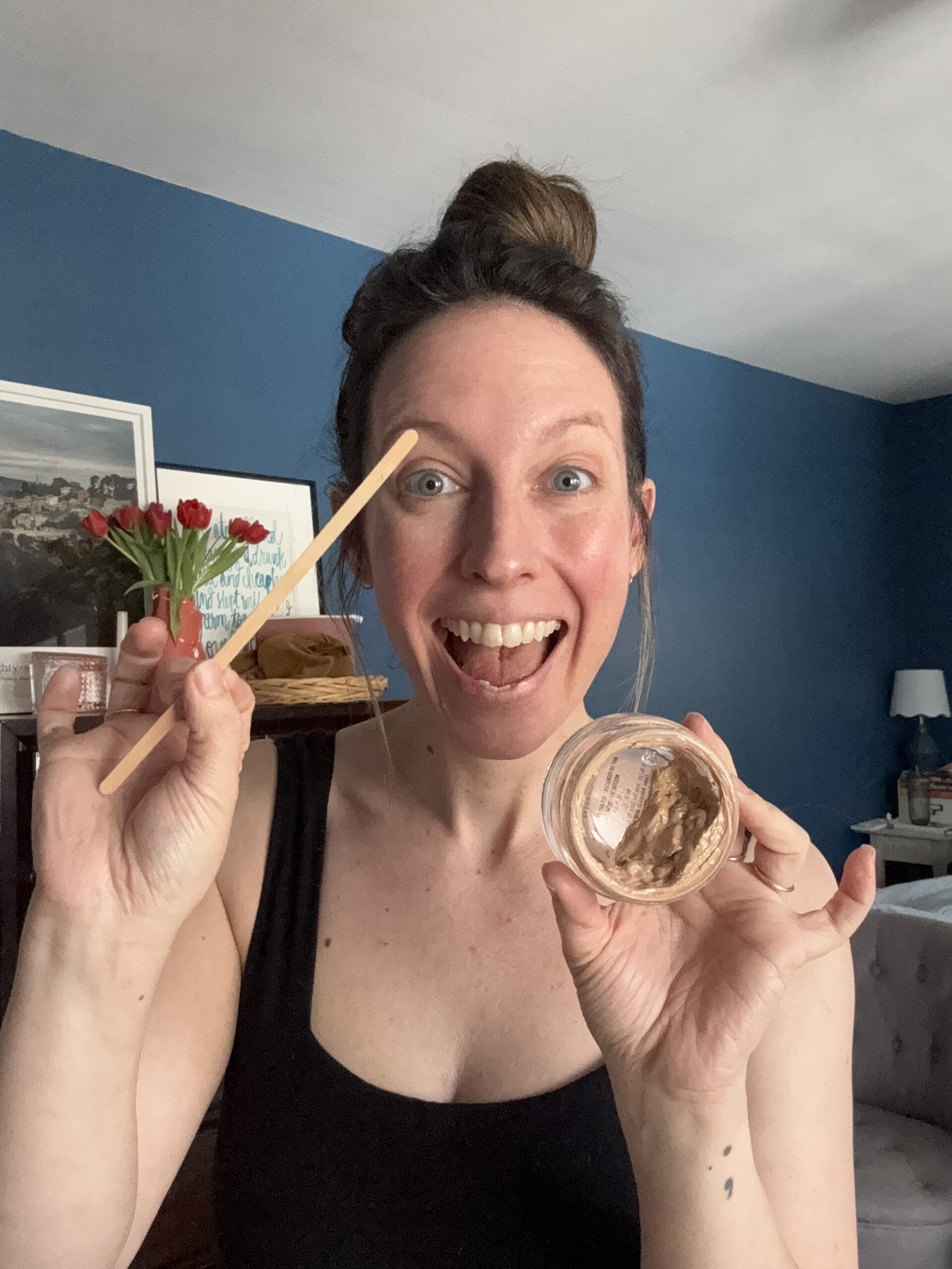 A woman in a black tank top smiles warmly, holding an empty container in one hand and a wooden stick in the other.