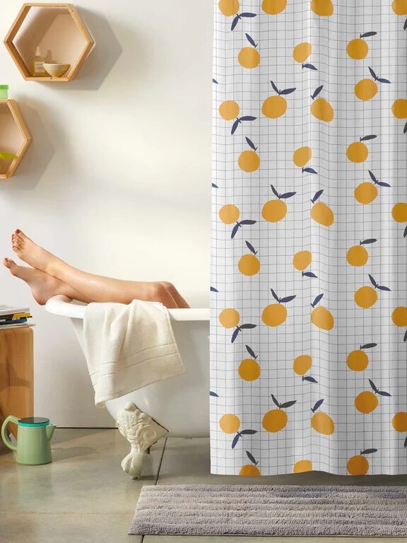 A person is relaxing in a bathtub behind a shower curtain decorated with orange fruit patterns. Only the person's legs are visible. The bathroom has modern decor elements, such as wall shelves and a mat.