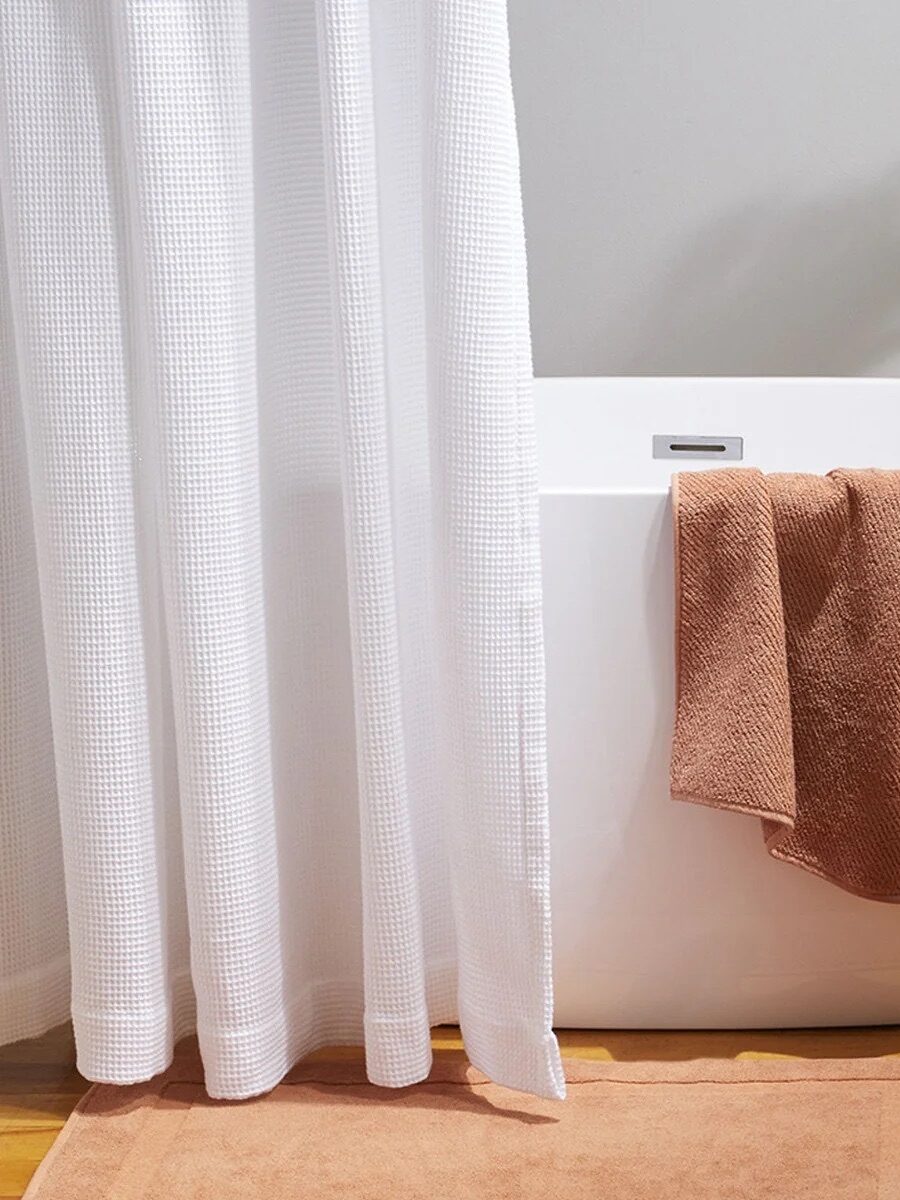 A white bathtub with a brown towel draped over the edge, a stack of brown towels on a wooden stool, a white shower curtain, and a green plant in the background.
