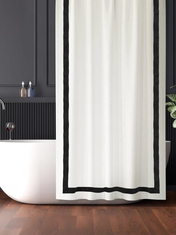 Modern bathroom with a freestanding white tub, black fixtures, and a white shower curtain with a black border. The dark gray walls feature vertical paneling, and a potted plant is in the corner.