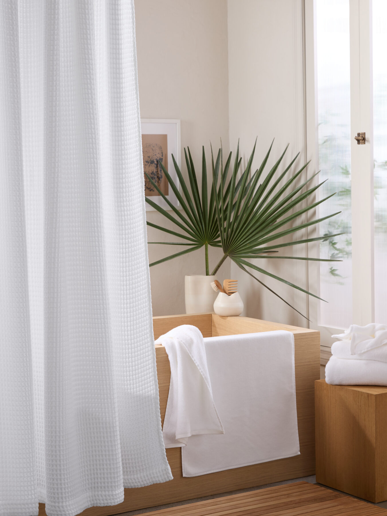 Clean, modern bathroom with a white shower curtain, a neatly folded towel on an open wood cabinet, and a plant next to the bathtub. Towels hang on adjacent wall hooks. Natural light streams in.