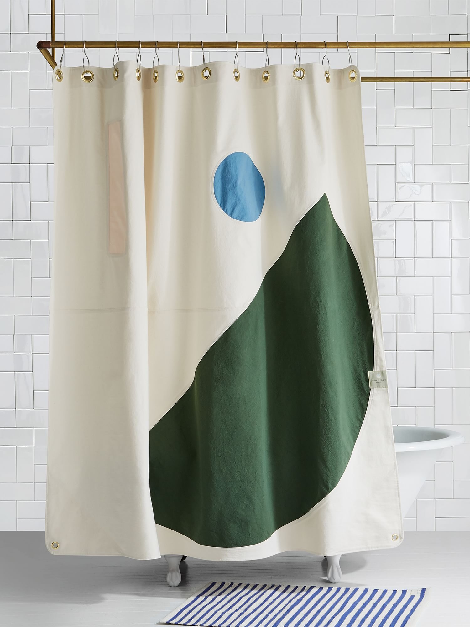 A shower curtain featuring geometric shapes in green and blue hangs in a bathroom with white tiled walls and a striped rug on the floor.