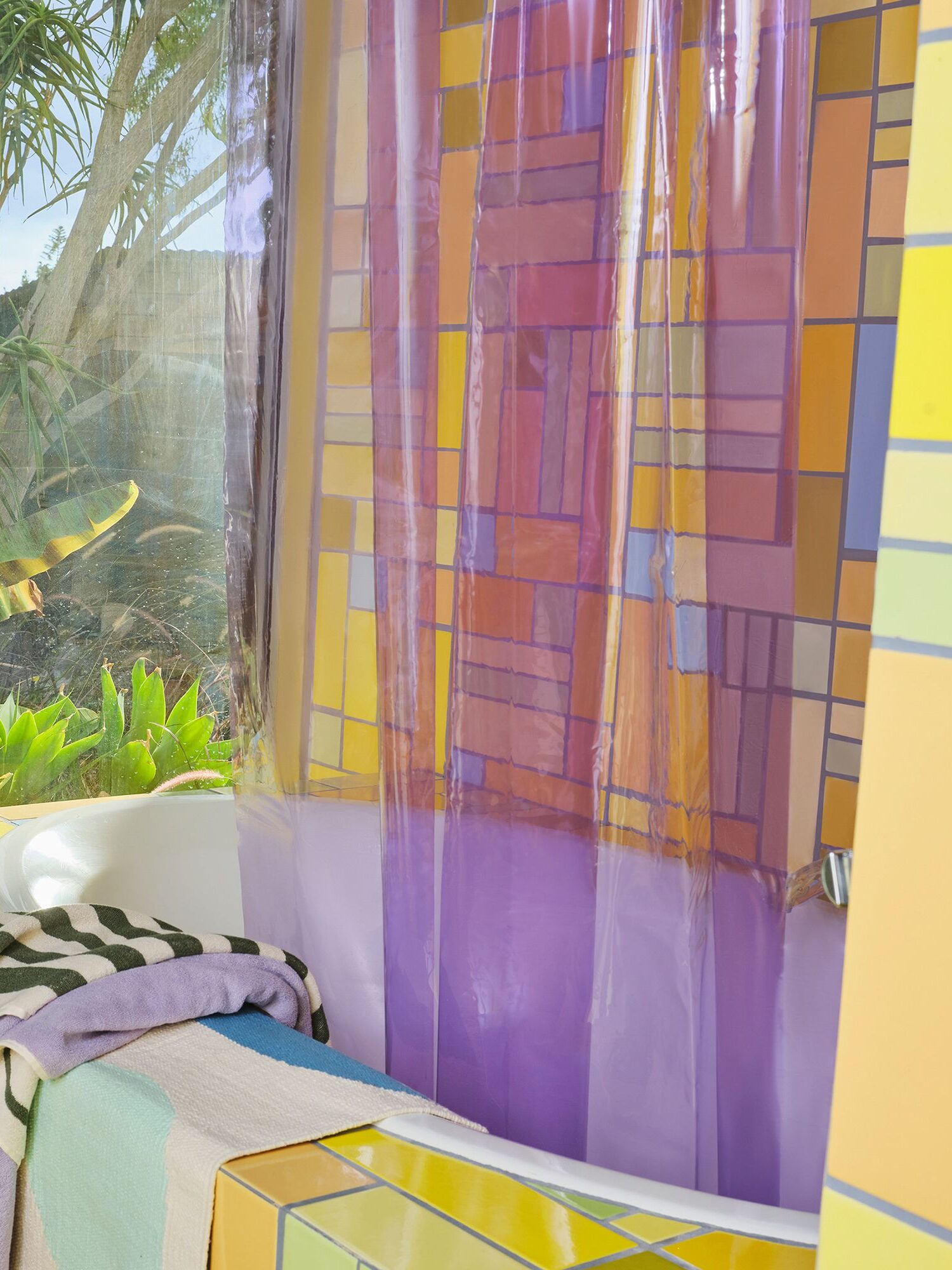 A colorful bathroom with a patterned tile design and a transparent shower curtain. The window reveals greenery outside. Towels are draped over a yellow tiled bench.