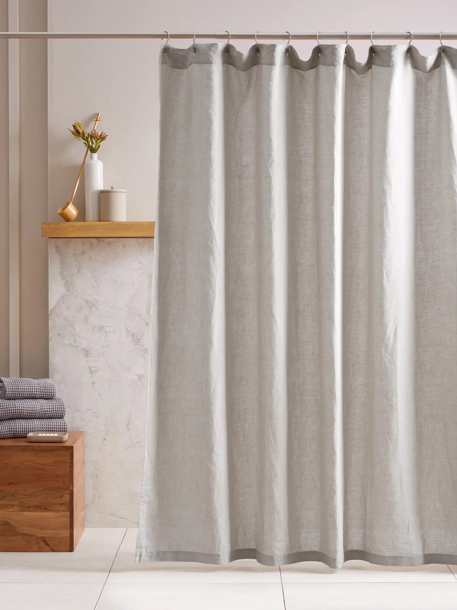 A minimalist bathroom features a light gray shower curtain, wooden bench with stacked towels, a shelf with a potted plant, and a beige wall with white tiles.