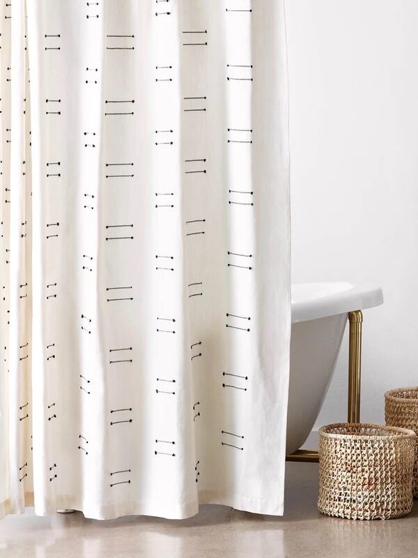 A bathroom with a white curtain featuring a modern linear pattern, a freestanding bathtub, two woven baskets on the floor, and a wooden ladder rack holding folded towels.