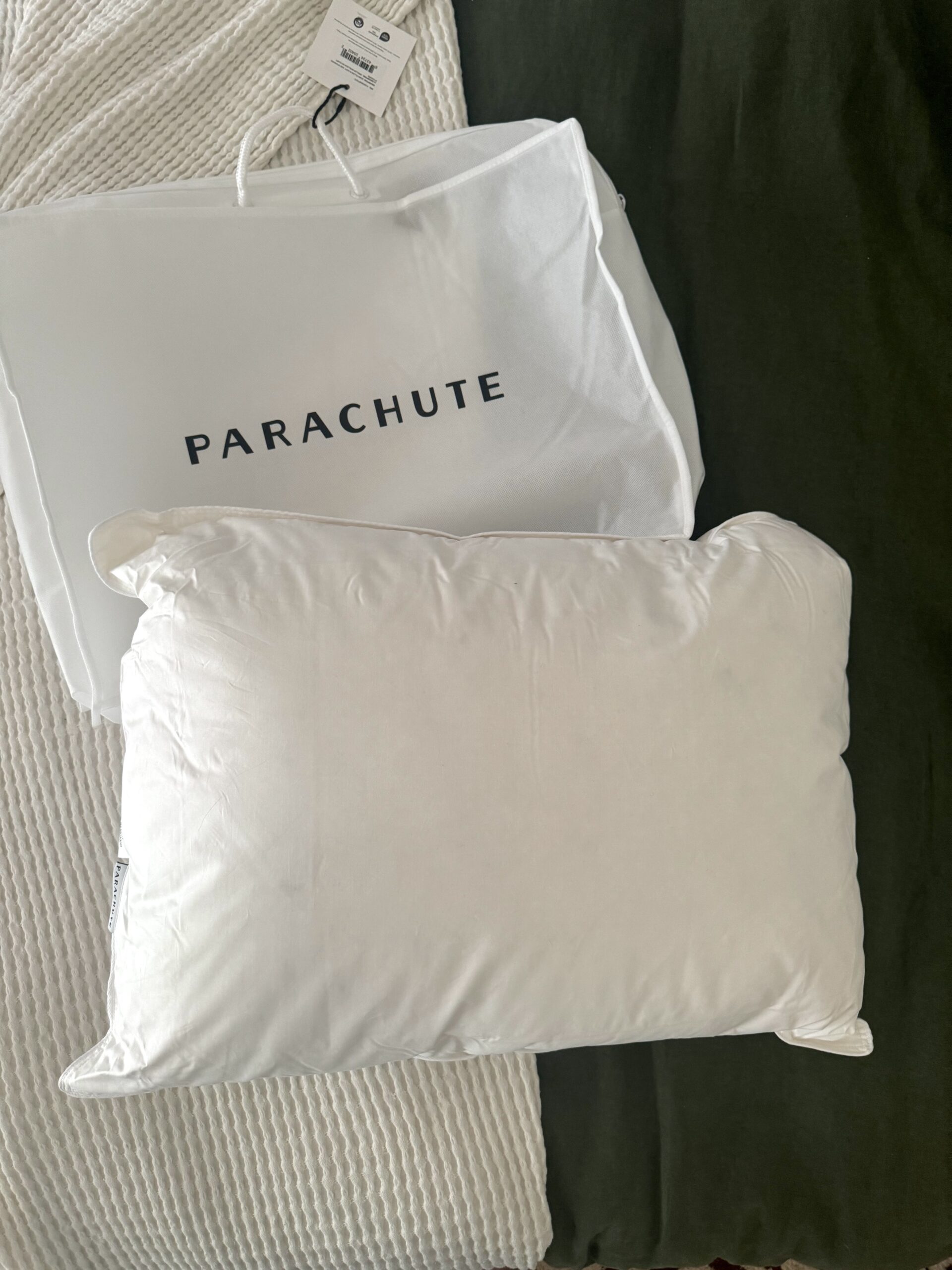 A white Parachute pillow with its matching branded protective bag is placed on a bed with a white and dark green blanket.