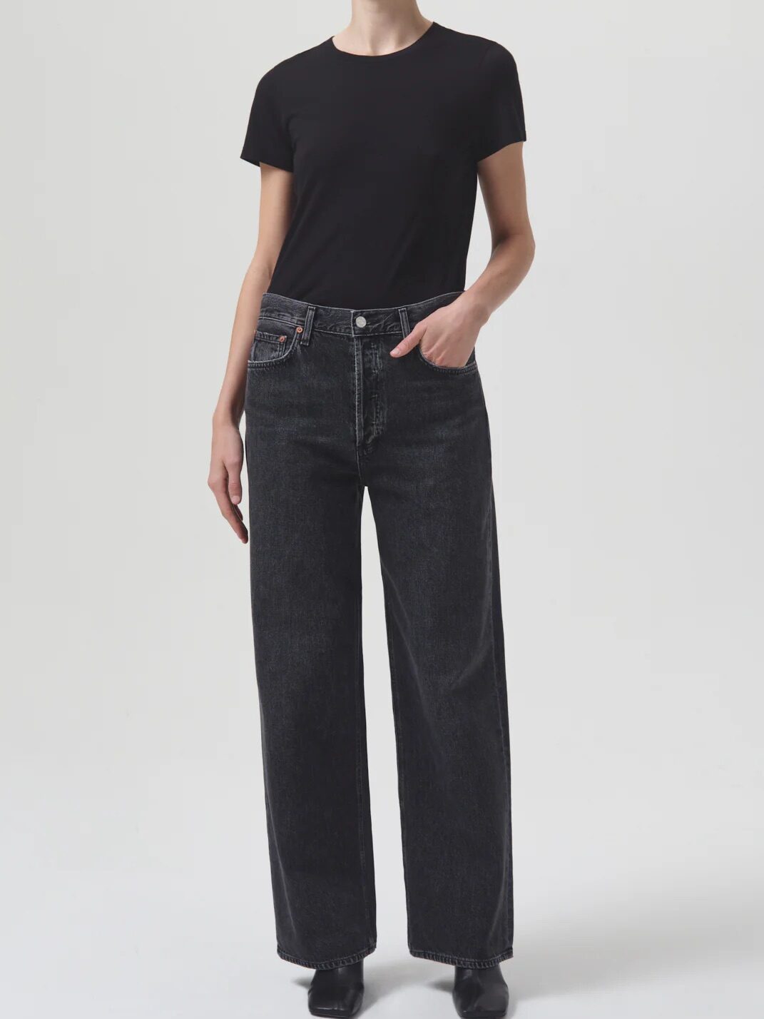 A person stands against a plain background wearing a black t-shirt and high-waisted, wide-leg black jeans. One hand is in their pocket.