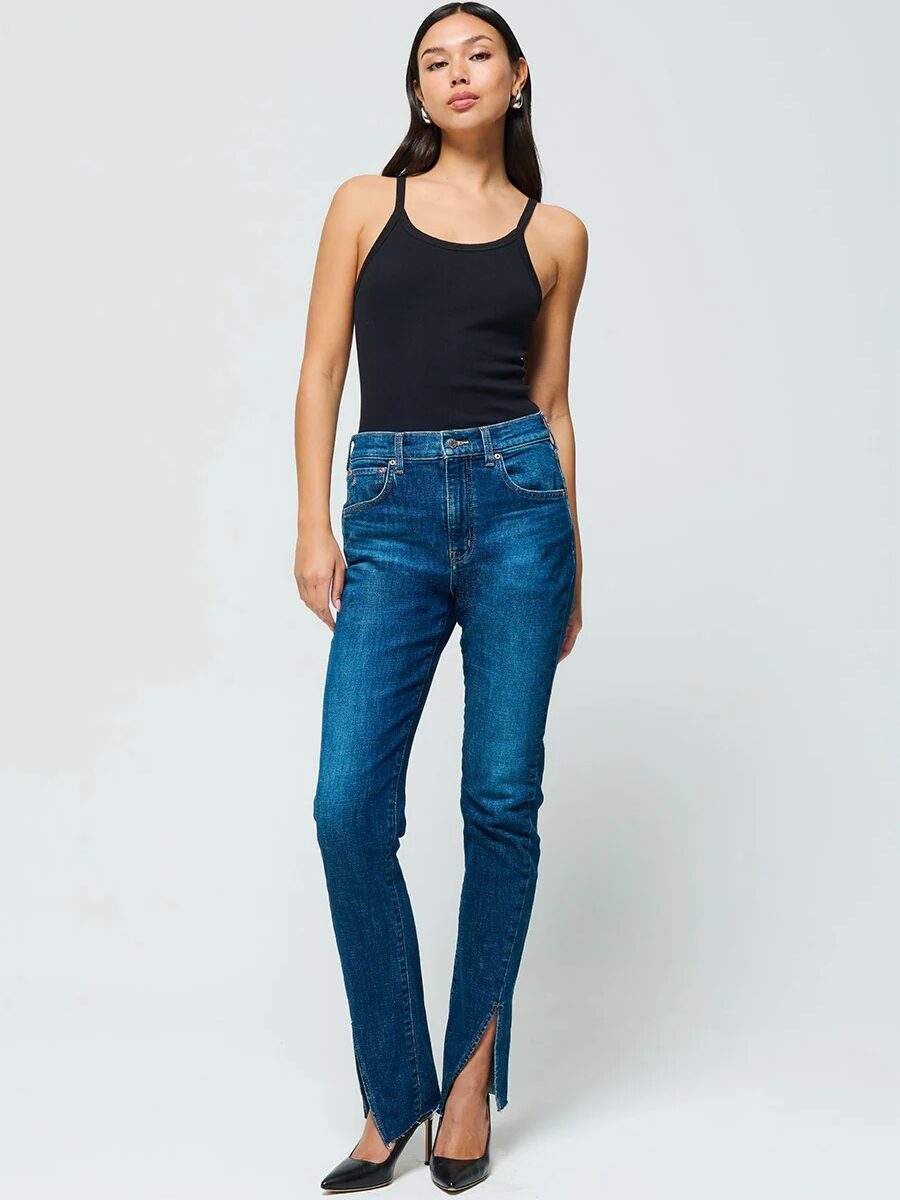 A woman stands against a white background wearing a black sleeveless top, blue jeans with slit hems, and black high-heeled shoes.