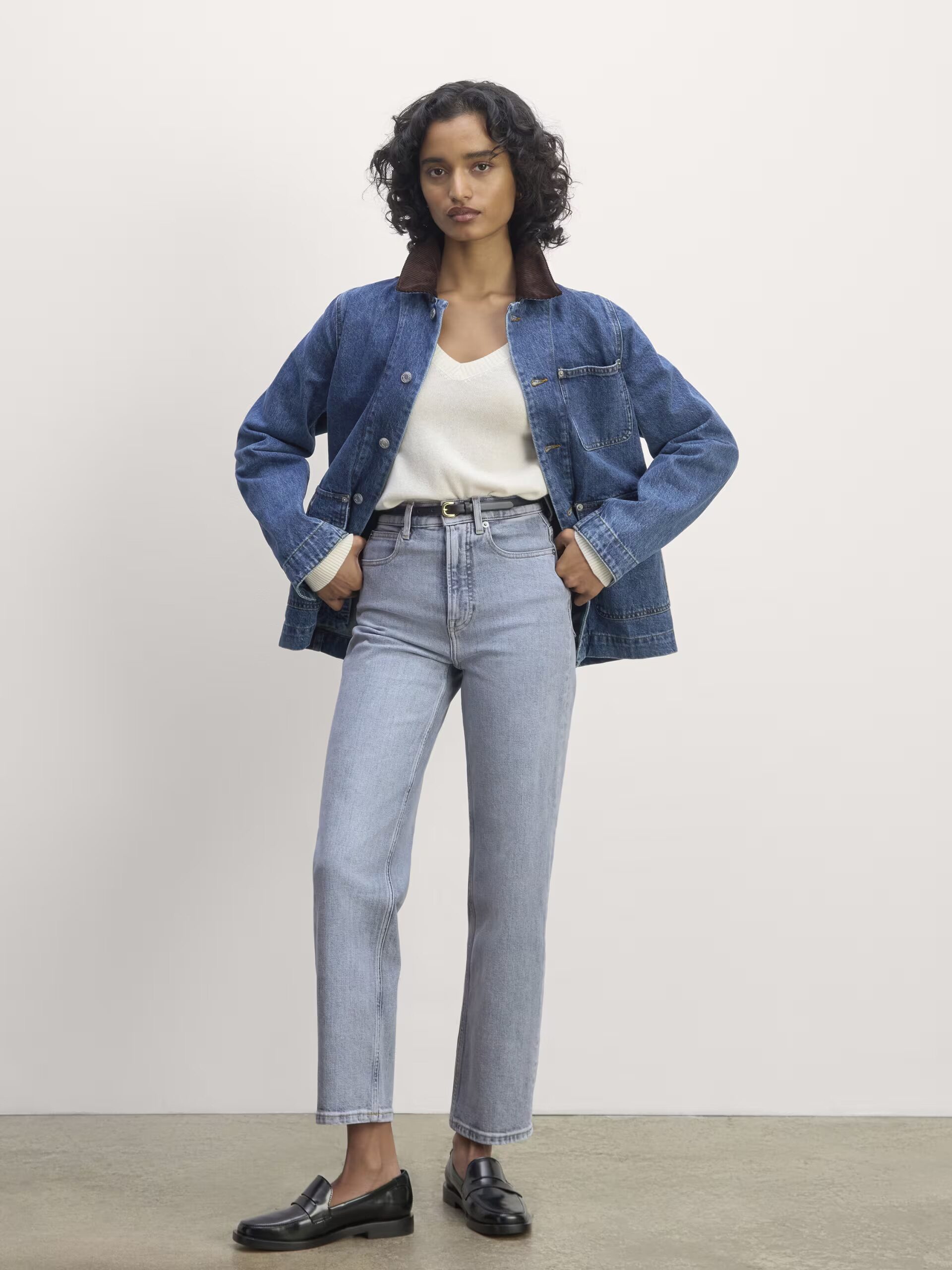 Person in a white V-neck top, blue denim jacket, light wash jeans, and black loafers standing against a plain white background.