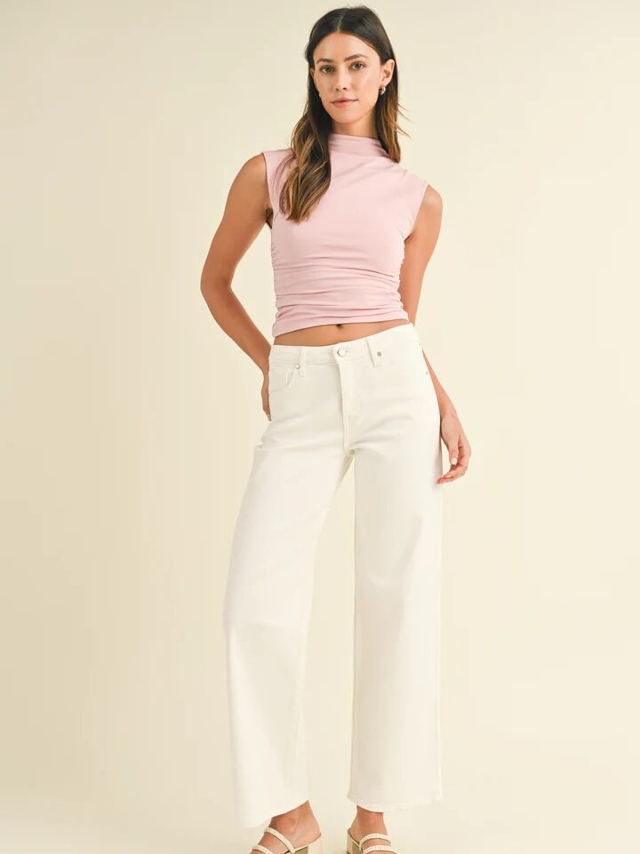 A woman stands against a plain background, wearing a sleeveless pink top, white wide-leg pants, and beige sandals. She has long brown hair and is posing with one hand on her hip.