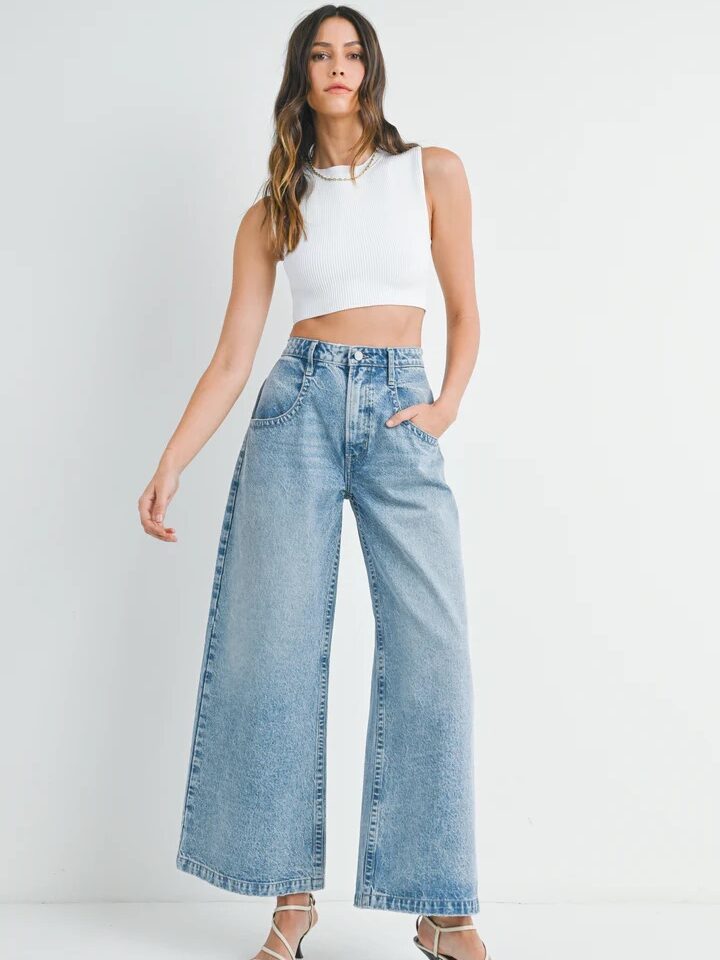 Woman stands against a plain background wearing a white sleeveless crop top paired with wide-leg denim jeans and strappy heels. She has one hand in her pocket.