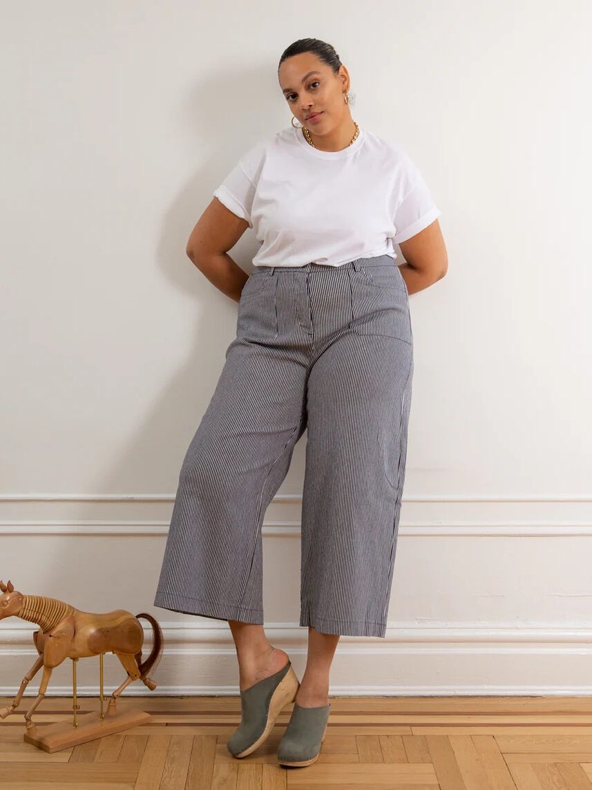 A person stands against a white wall and wooden floor, wearing a white t-shirt, gray wide-leg pants, and green shoes, with a wooden toy horse positioned on the floor next to them.