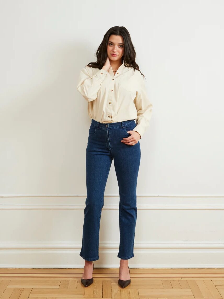 A woman stands against a white wall, wearing a beige button-up shirt, blue jeans, and brown shoes. She has one hand on her neck and the other hand by her side.