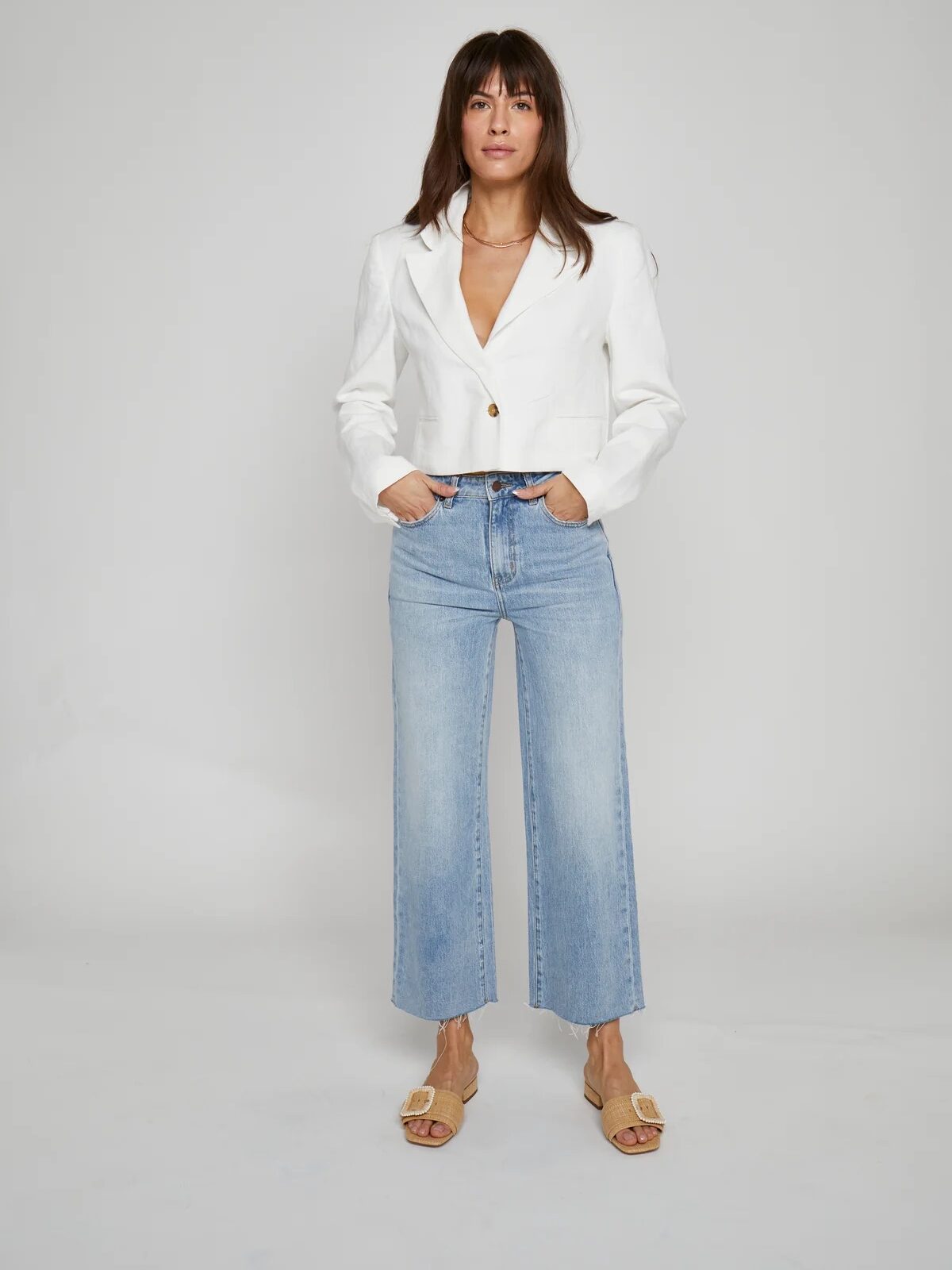 A woman stands against a plain background wearing a white blazer, light blue high-waisted jeans, and beige sandals.