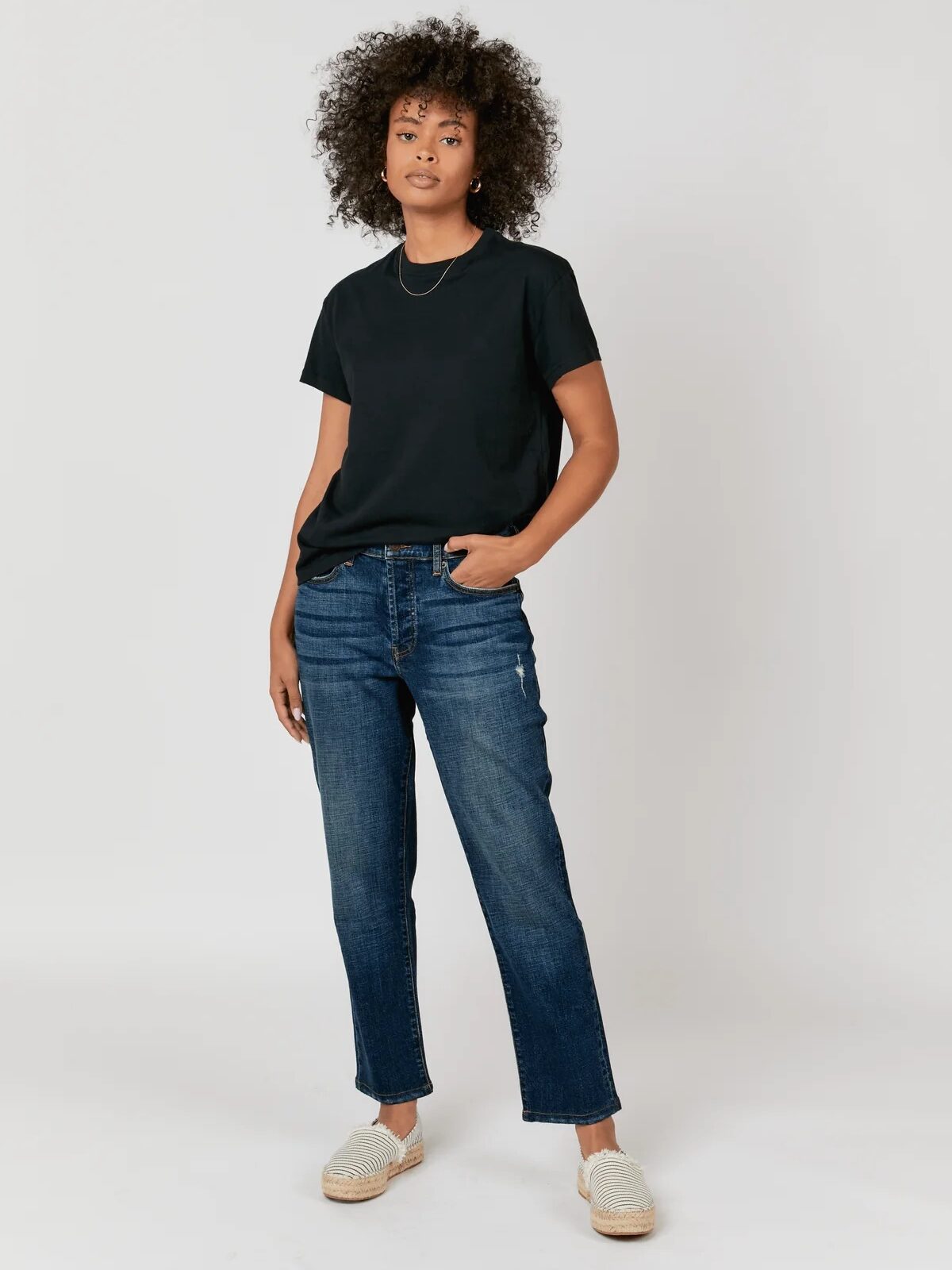 Person wearing a black t-shirt, blue jeans, and white slip-on shoes stands against a plain white background.