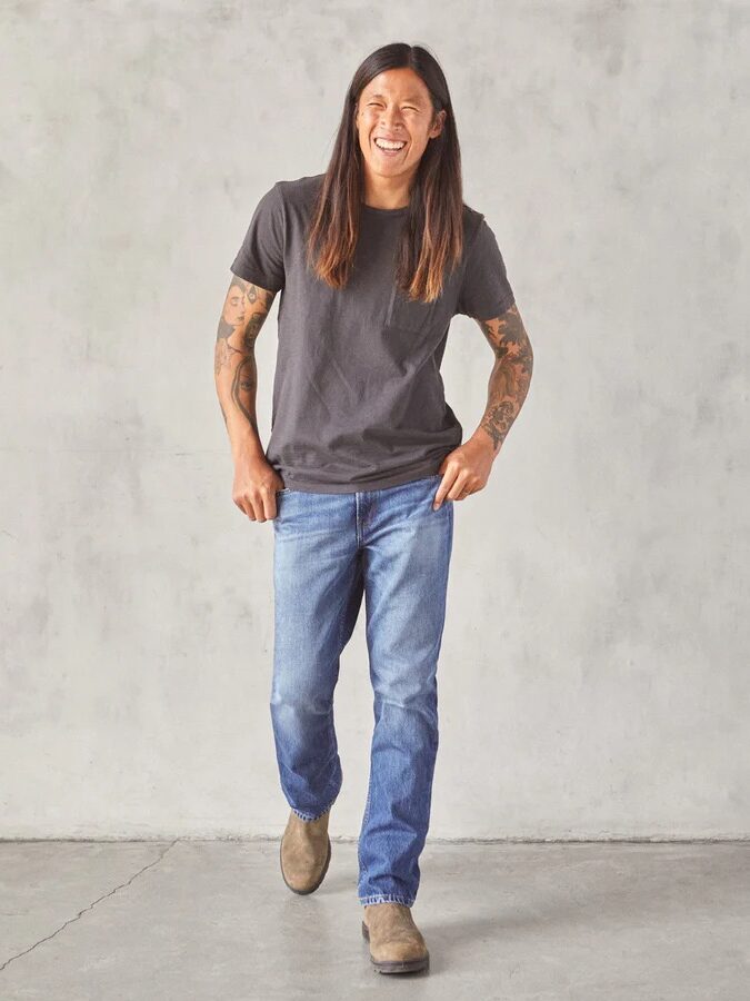 A person with long hair, wearing a grey t-shirt, blue jeans, and brown shoes, is walking and smiling against a plain grey wall. Their arms are tattooed.
