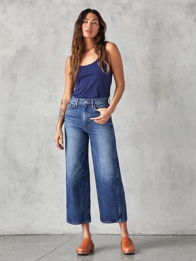 A woman with long hair stands against a plain, textured wall. She is wearing a navy tank top, high-waisted wide-leg jeans, and brown slip-on shoes.