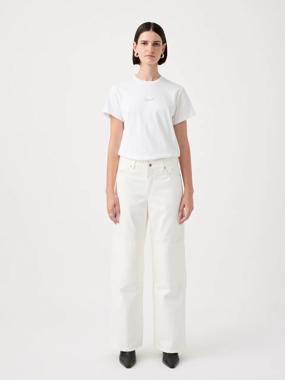 A person stands facing forward, wearing a white T-shirt, white high-waisted wide-leg jeans, and black pointed shoes, against a plain white background.