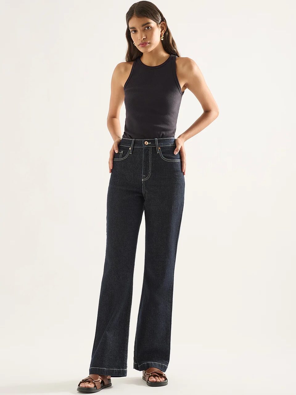 A person stands posing against a light background, wearing a black sleeveless top, high-waisted dark blue jeans, and black sandals.