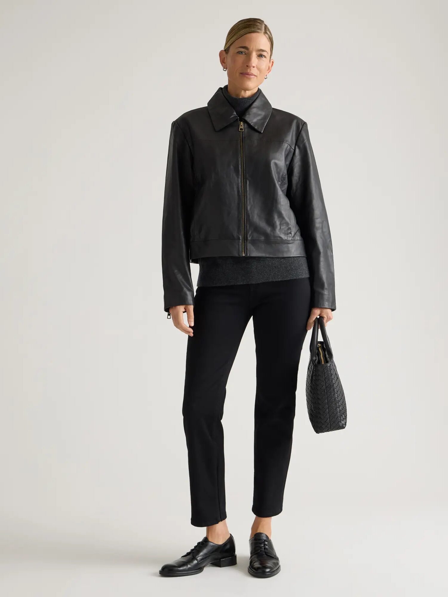 A person stands against a plain background wearing a black leather jacket, dark pants, and black shoes, holding a textured black handbag.