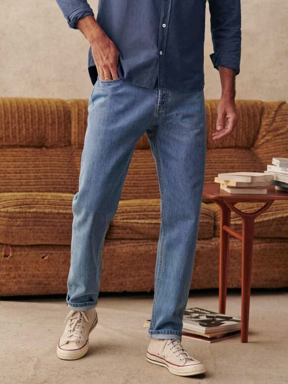 A person stands in front of a brown couch wearing a blue shirt, light blue jeans, and white sneakers. One hand is in a pocket, and several books are stacked on a wooden side table.