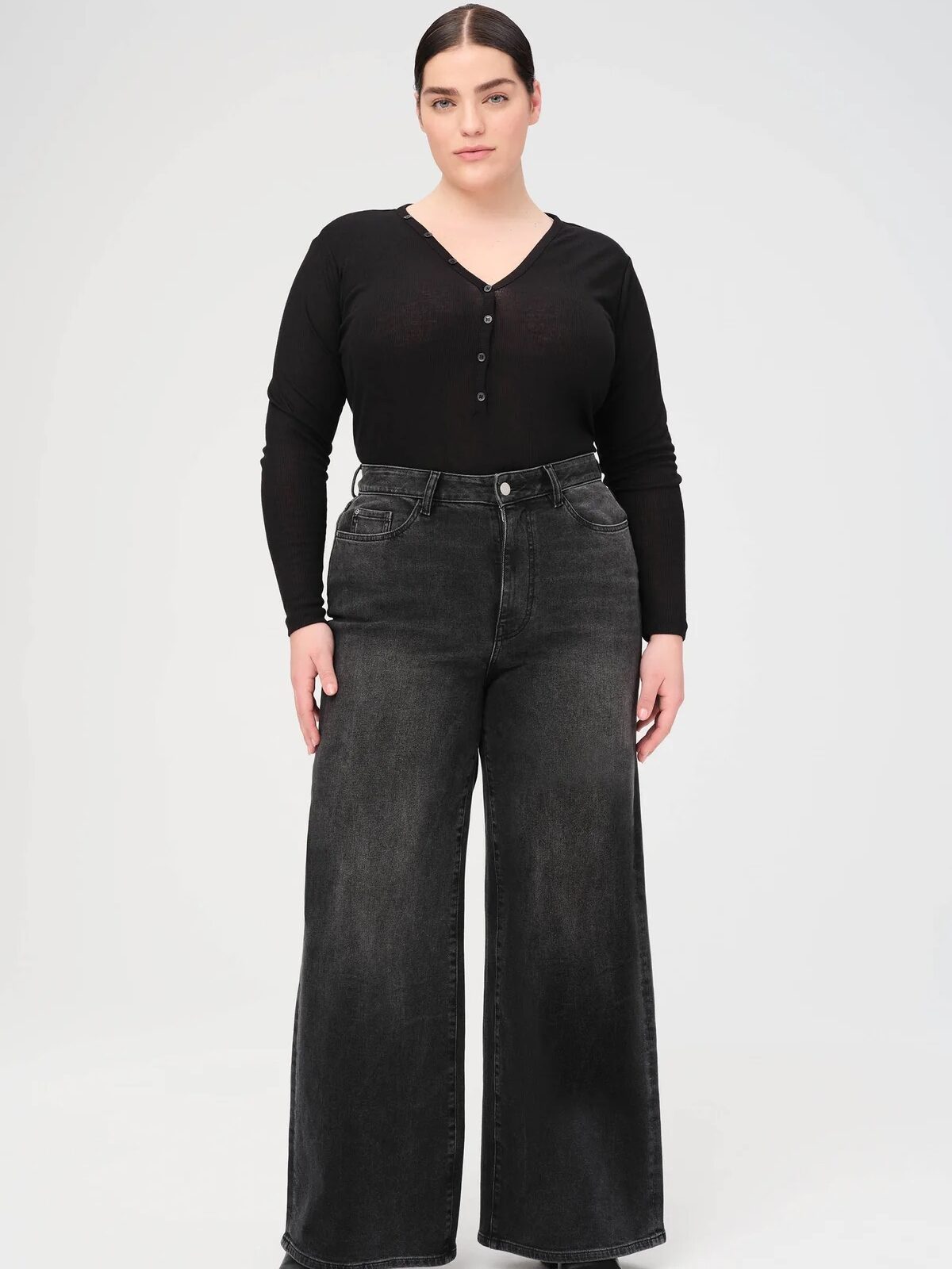 A woman stands with a neutral expression, wearing a black long-sleeve top and high-waisted wide-leg jeans. She has dark hair pulled back and is in front of a plain white background.