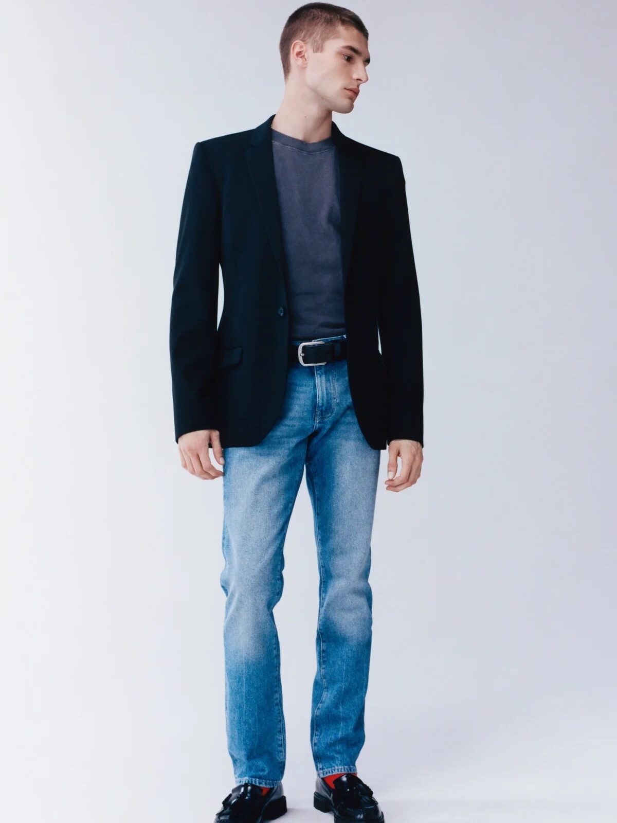 A person wearing a black blazer, a dark grey t-shirt, denim jeans, and black shoes stands against a light background.