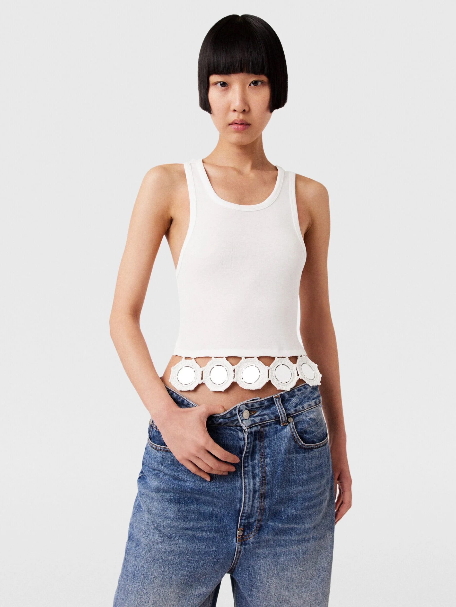 Person wearing a white sleeveless top with circular cutouts at the hem and blue jeans, standing against a plain background.