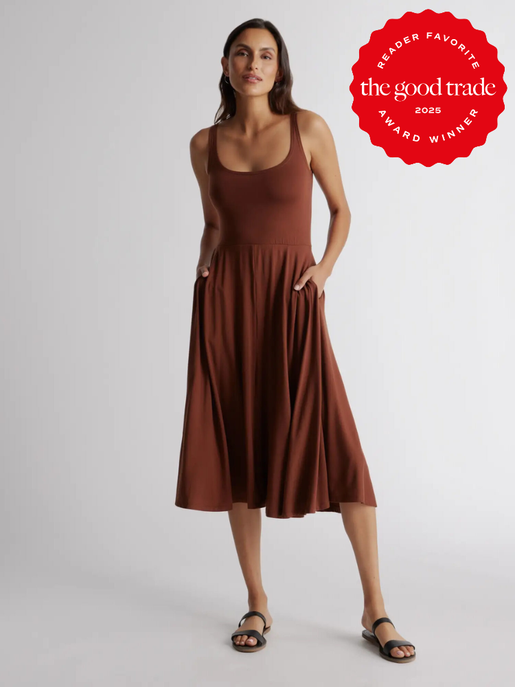 Person in a brown sleeveless dress stands with hands in pockets. The dress has a flared skirt. There is an award badge in the top right corner.