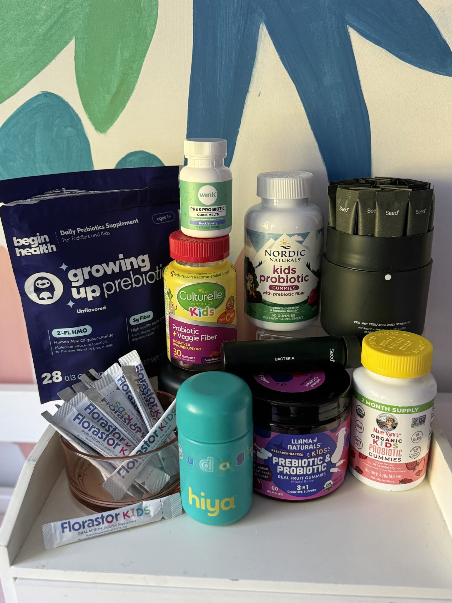 A variety of kids’ probiotic and prebiotic supplements, including powders, chewables, and gummies, are displayed on a white shelf in front of a colorful painted wall.
