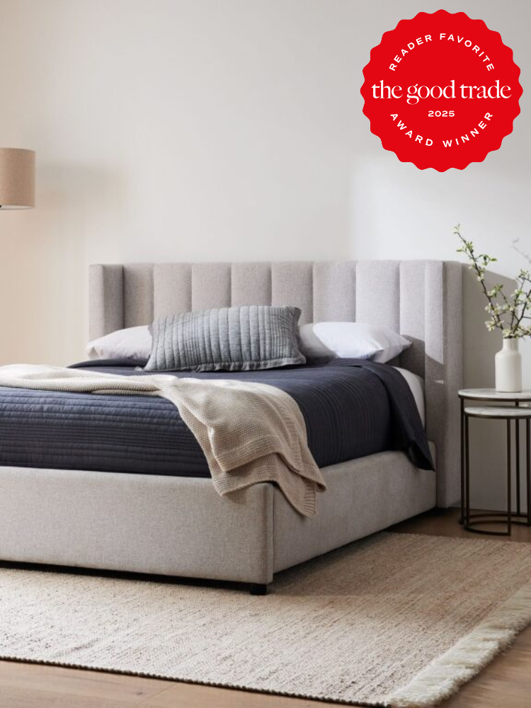 A modern bedroom with a gray upholstered bed, striped gray pillow, and layered blankets. A white lamp and vase with branches are on a nightstand. "The Good Trade 2025 Award Winner" badge in corner.