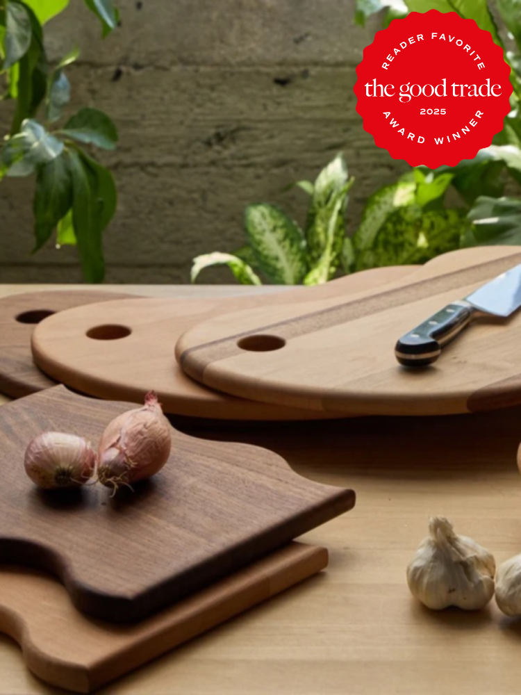 Wooden cutting boards with a knife and garlic on a table; green plants in the background. "The Good Trade 2025 Award Winner" badge in the top right corner.