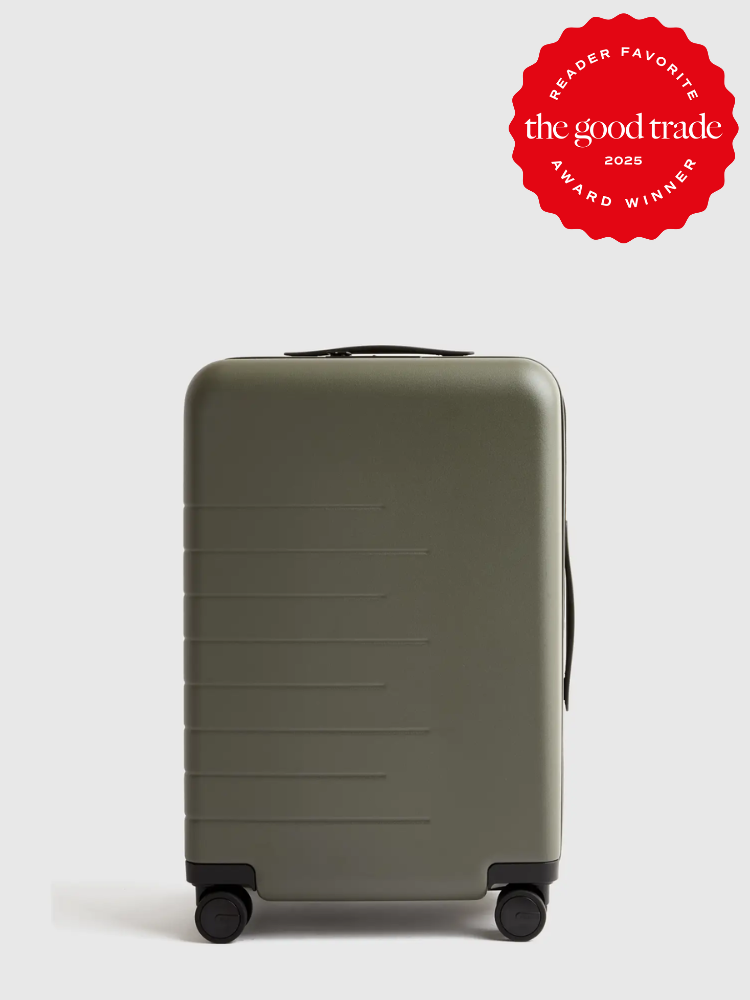 A dark green hard-shell suitcase with four wheels and a side handle, featuring a "Reader Favorite: The Good Trade Award Winner 2025" red badge in the top right corner.
