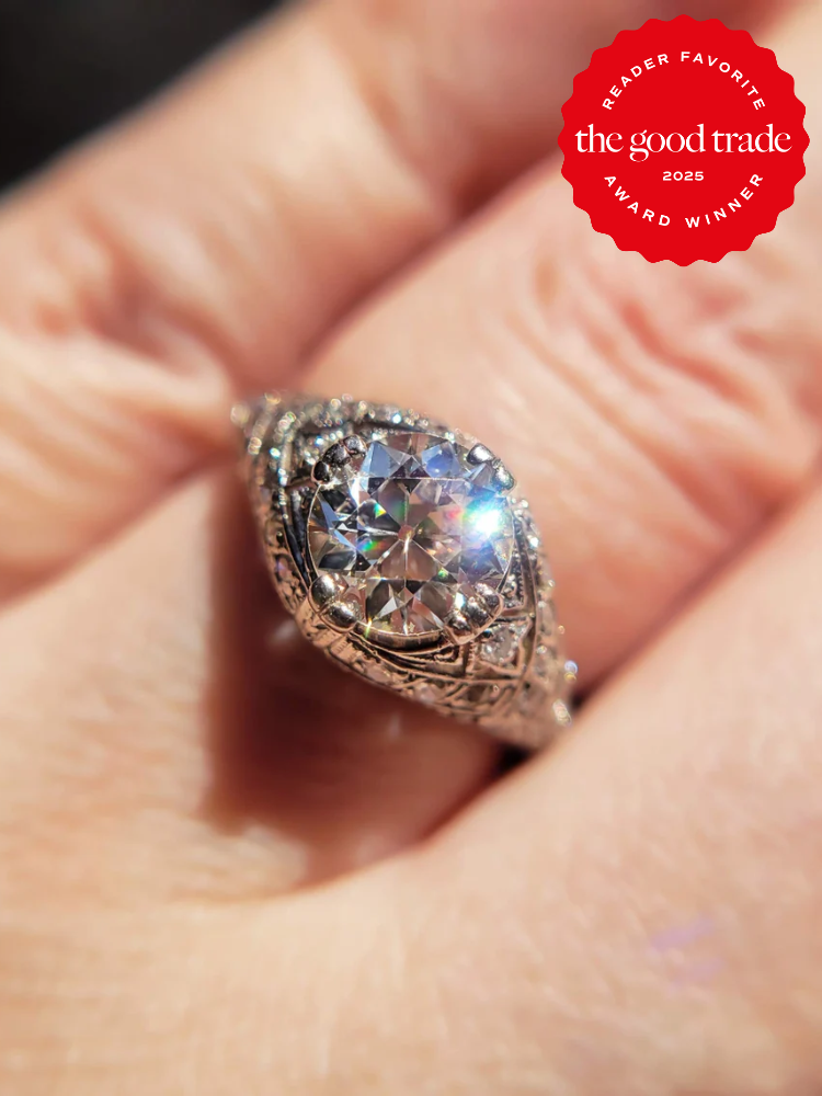 Antique-style ring with a large, sparkling center stone and intricate details, worn on a finger. Red "The Good Trade 2025 Reader Favorite Award Winner" badge in the corner.