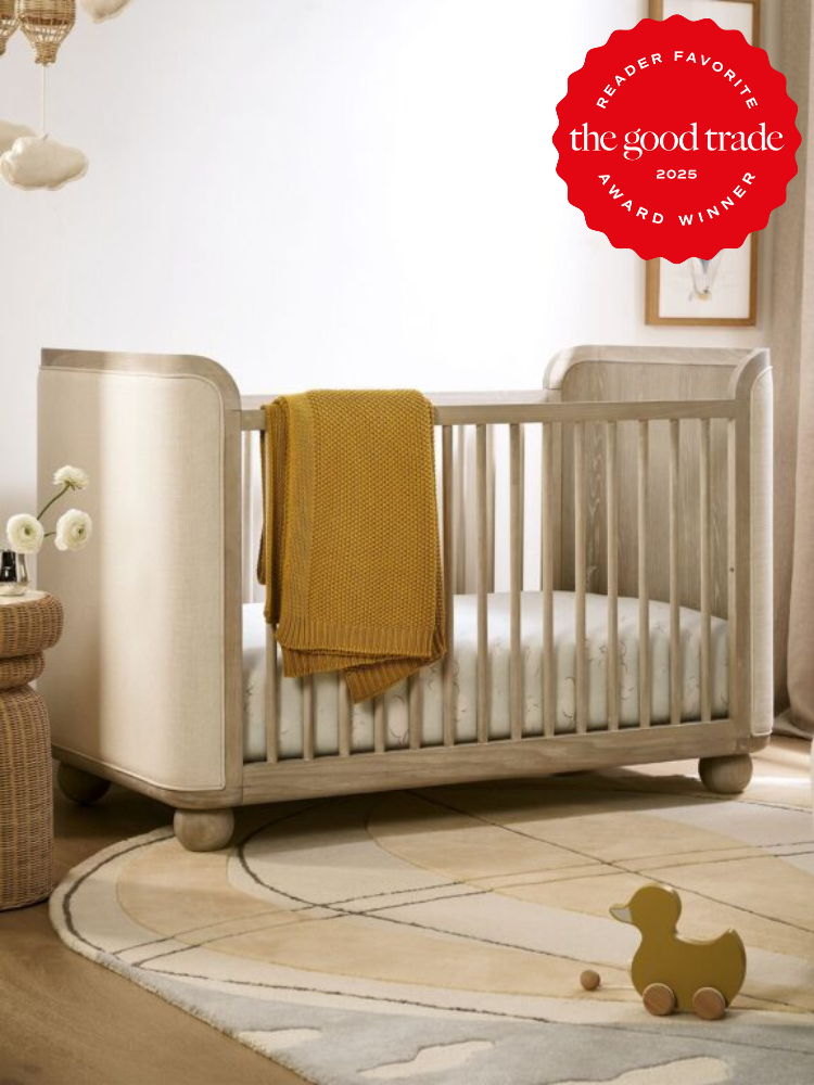 A wooden baby crib with a yellow blanket draped over the side, set on a circular rug. A small wooden duck toy sits nearby. A "2025 Reader Favorite The Good Trade Award Winner" badge is displayed.