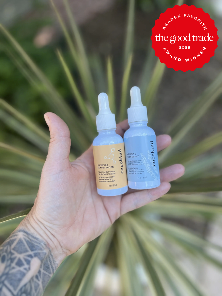 A tattooed hand holds two dropper bottles labeled "Cocokind," with a plant in the background. A red ribbon indicates a 2025 award from "The Good Trade.