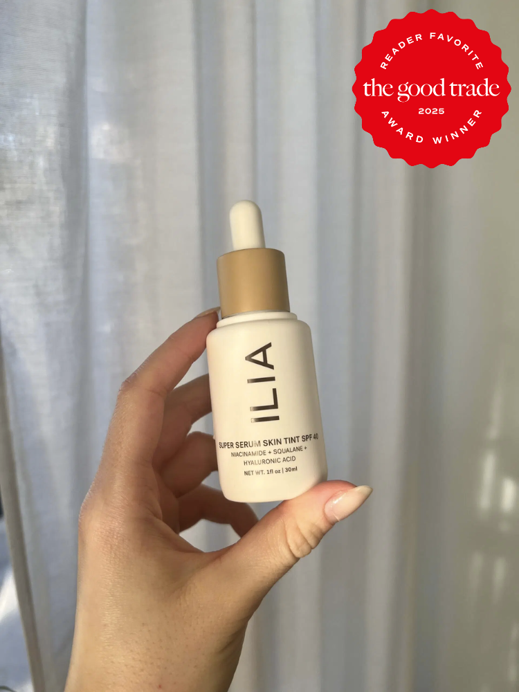 Hand holding a bottle of ILIA Super Serum Skin Tint SPF 40 against a neutral background, with a red badge stating "Reader Favorite, The Good Trade, Award Winner 2025.