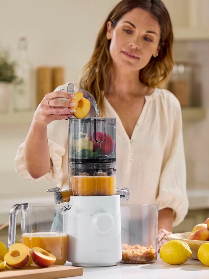 Woman using a juicer in a kitchen, adding peach slices. Fresh juice is in a container below, surrounded by peaches on the counter.