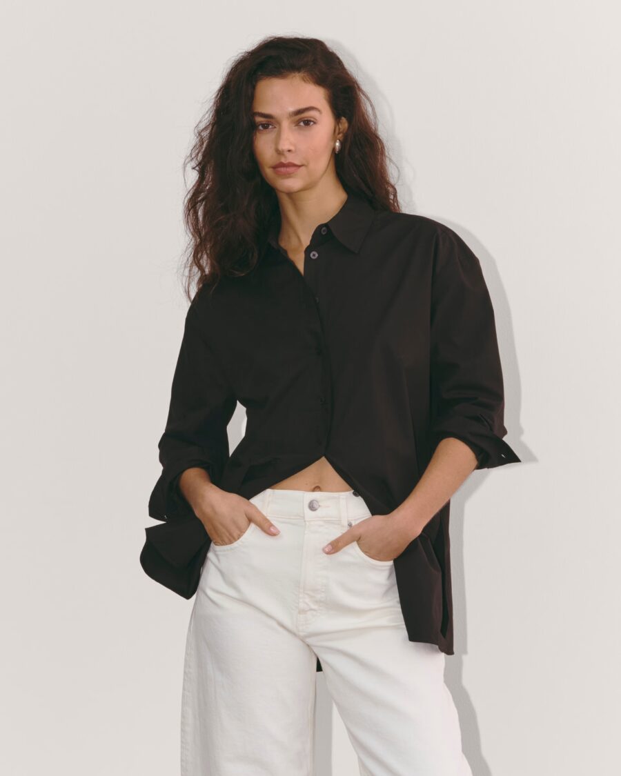 A person with long hair is wearing a black button-up shirt and white pants, standing against a plain background.