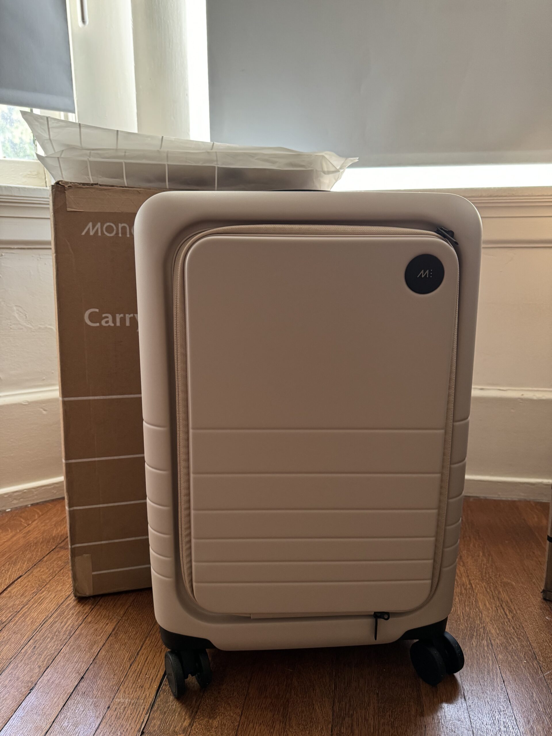 A beige hard-shell carry-on suitcase with four wheels is placed in front of a cardboard box on a wooden floor near a window.