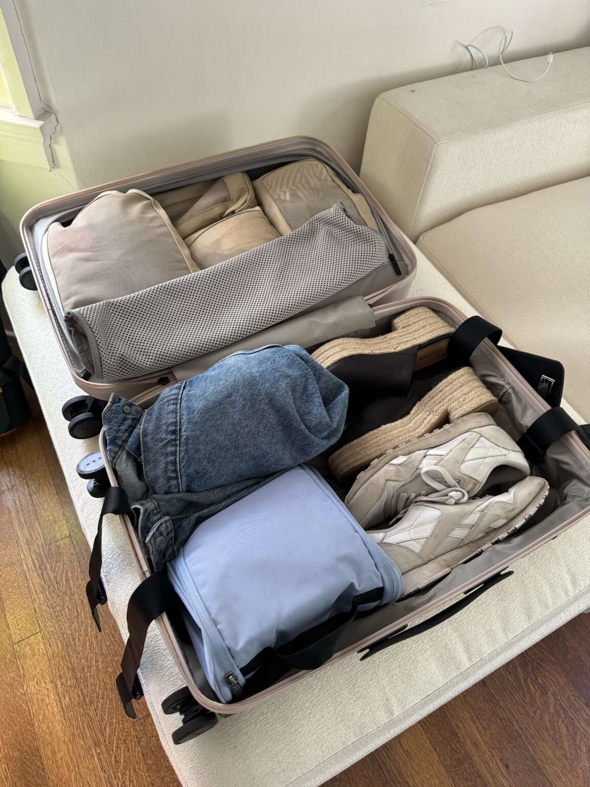 An open suitcase on a bed contains folded clothes, a pair of sneakers, sandals, a denim hat, and a light blue packing cube.