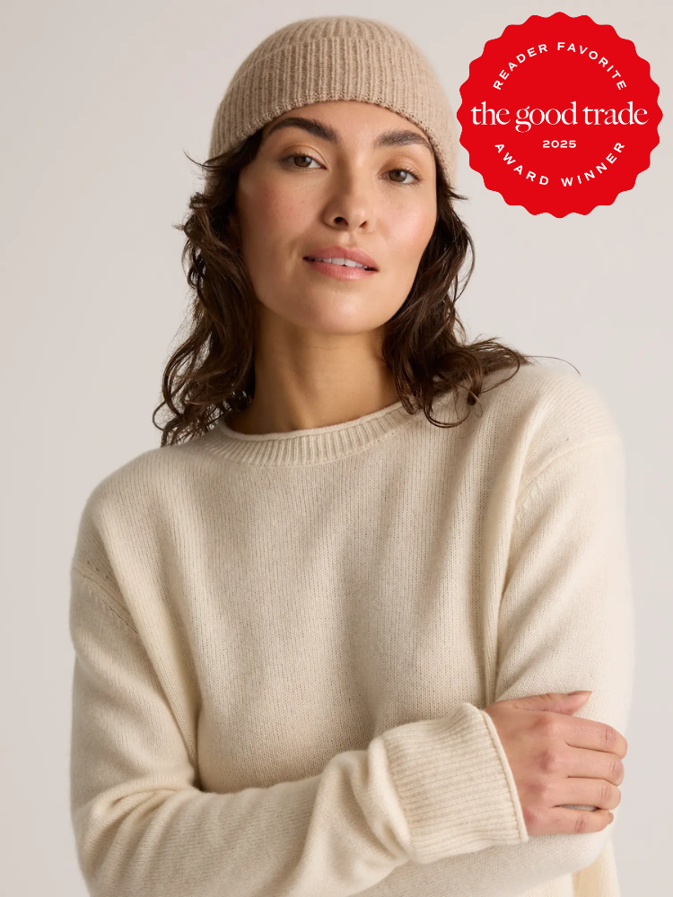 A woman wearing a beige knit beanie and cream sweater stands against a neutral background. A red badge in the corner reads "The Good Trade 2025 Award Winner.