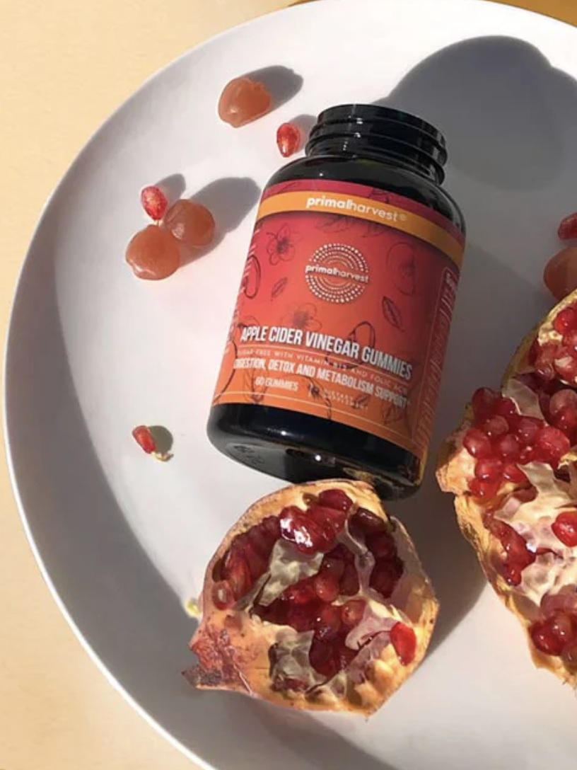 A bottle of apple cider vinegar gummies on a plate with a cut pomegranate and a few loose gummies.