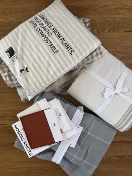 A collection of folded textiles, including blankets and swatches, lies on a wooden floor. A labeled package reads "Made from plants. No plastic. 100% compostable." Swatches have tags marked "NK.