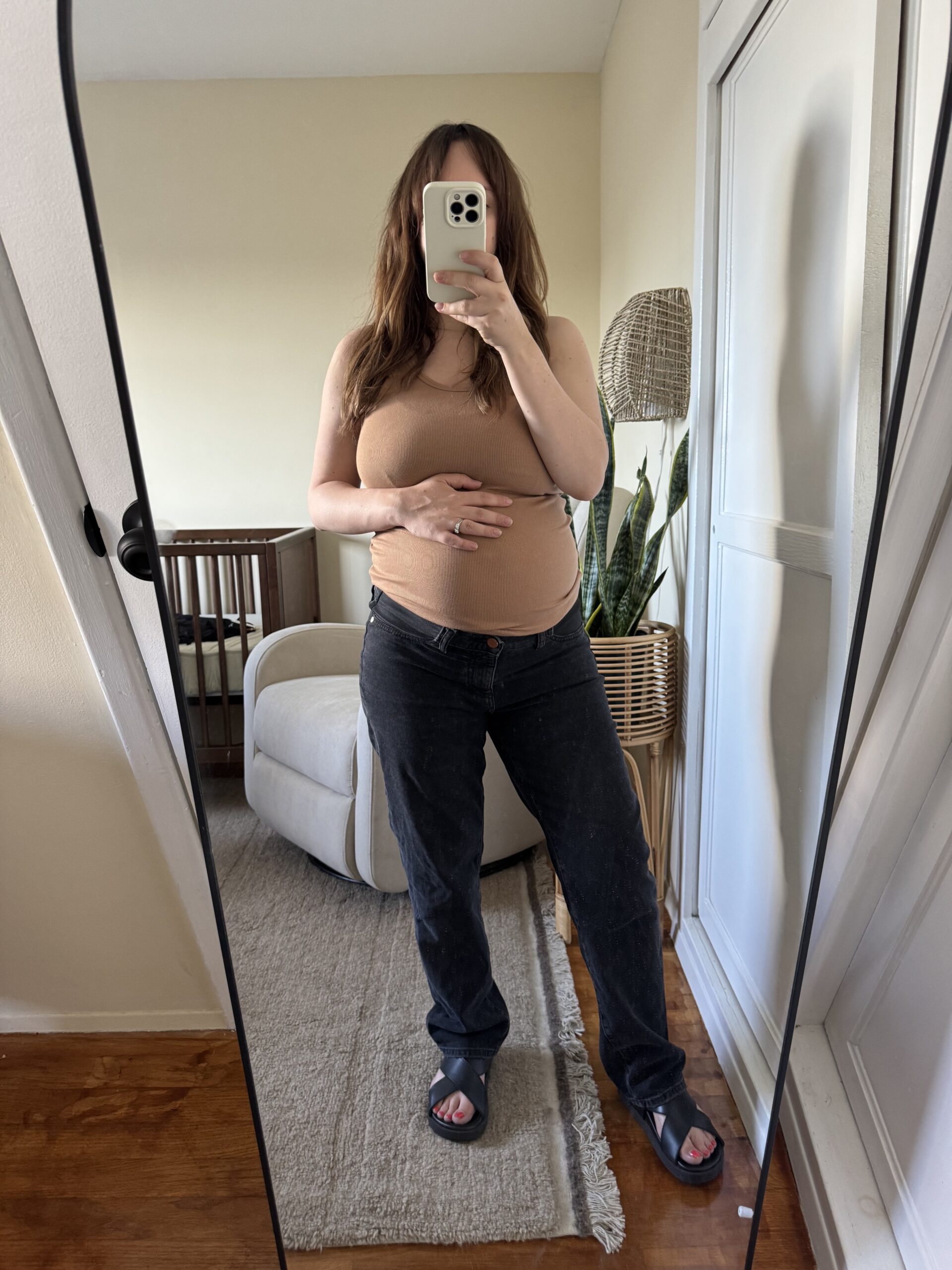 A person stands in front of a mirror, holding their phone and their stomach, wearing a beige tank top, black pants, and sandals in a softly lit room with neutral decor.