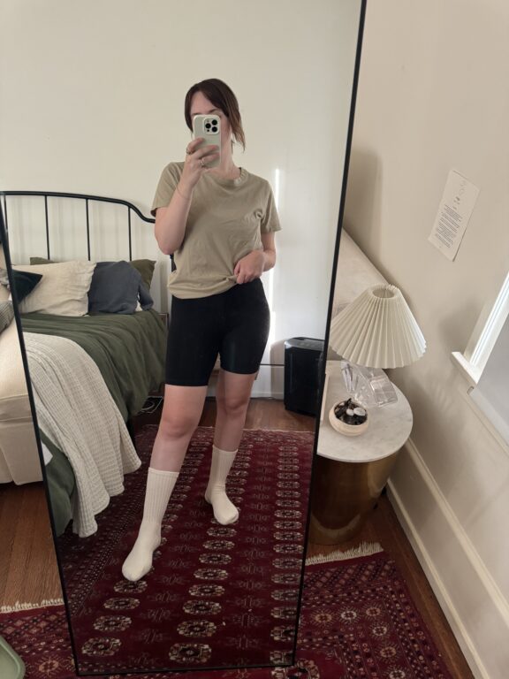 Person taking a mirror selfie in a bedroom, wearing a beige t-shirt, black shorts, and white socks. The room has a bed, nightstand, lamp, and a red patterned rug.