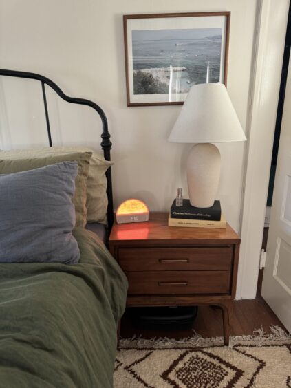 A neatly made bed with green bedding is next to a wooden nightstand holding a lamp, a digital clock with an orange glow, two books, and a framed photo on the wall above.