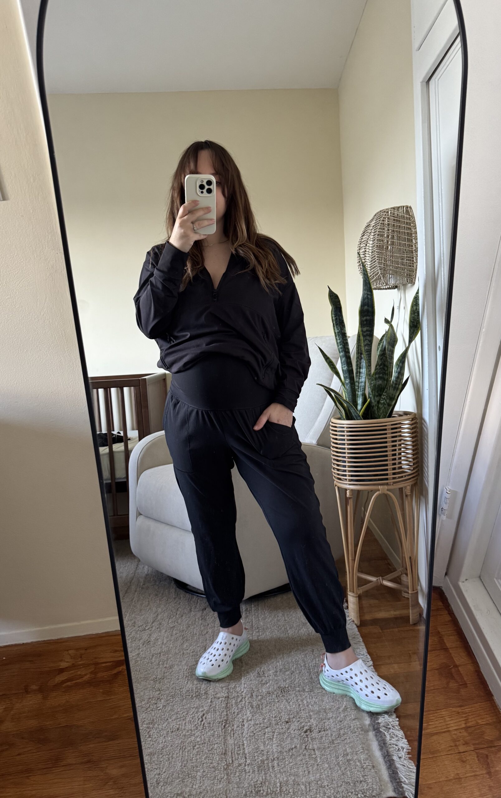 Person taking a mirror selfie in a room, wearing a black outfit with white and green clogs. There is a white armchair, a crib, and a potted plant in the background.