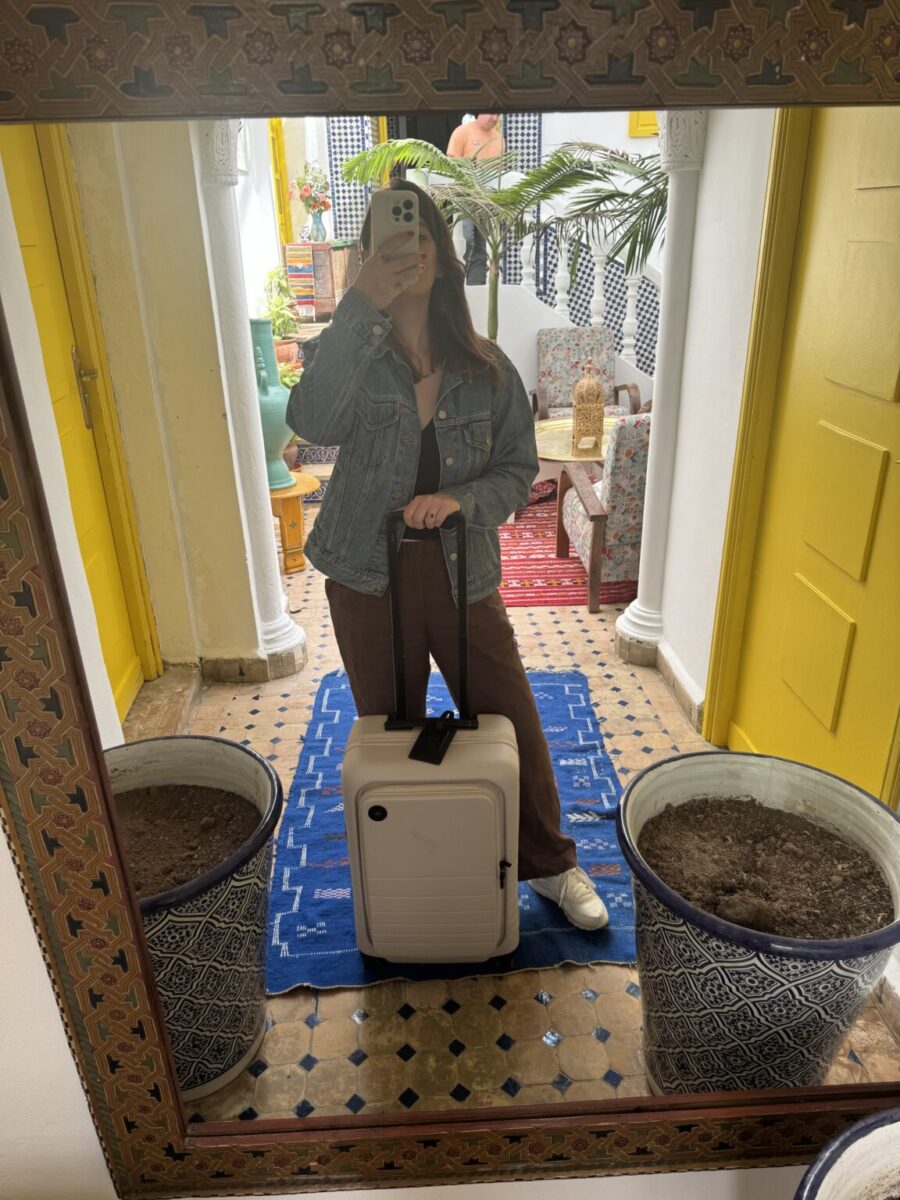 Woman takes a mirror selfie in a bright room, standing behind a white suitcase. She wears a denim jacket and brown pants. The room has yellow doors, blue decor, and potted plants.