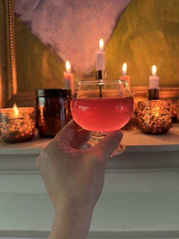 A hand holds a glass of pink liquid in front of lit candles on a mantel, with a framed painting in the background.